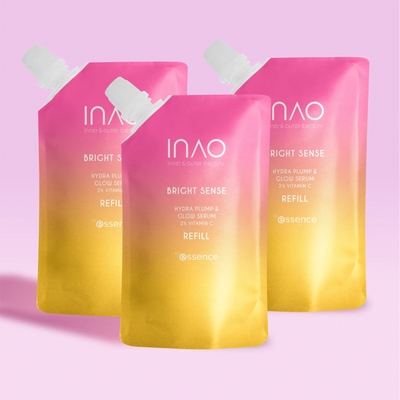 Drei rosa und gelbe Farbverlaufs-Nachfüllbeutel des INAO by essence Bright Sense Hydra Plump & Glow Serum Refill mit 2 % Vitamin C und Hyaluronsäure stehen aufrecht vor einem hellrosa Hintergrund.