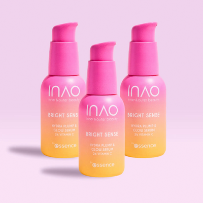 Drei rosa und gelbe Farbverlaufsflaschen des INAO by essence Bright Sense Hydra Plump & Glow Serums mit 2 % Vitamin C und Hyaluronsäure stehen aufrecht auf einem hellrosa Hintergrund und tragen jeweils unten das „essence“-Logo.