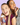 Zwei lächelnde Frauen in glänzenden Outfits posieren gemeinsam vor einem hellvioletten Hintergrund. Die eine hält eine Flasche „Clean Skin Community Edition Gummies“ von INAO by essence in der Hand, die andere ist gerade dabei, ein Gummibärchen zu essen.
