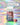 Eine Flasche INAO by essence Clean Skin Community Edition Gummies steht neben zwei kleinen Kristallen auf einem pastellfarben schimmernden Untergrund. Darüber verkünden deutsche Sprechblasen die erste Community Edition.