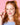 Eine lächelnde junge Frau mit roten Zöpfen zwinkert und hält ein kleines rosa Clean Skin x JUICY BOMB Gummies Bonbon von INAO by essence vor ihrem Gesicht vor einem rosa Hintergrund. Sie trägt ein hellblaues Top, rosa Lippenstift und eine Perlenkette.