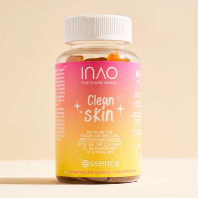 Eine Flasche INAO by essence Clean Skin Gummies mit weißem Verschluss enthält orangefarbene Gummibärchen. Das rosa-gelbe Etikett trägt weißen Text, und die Flasche steht auf einer beigen Fläche mit beigem Hintergrund.