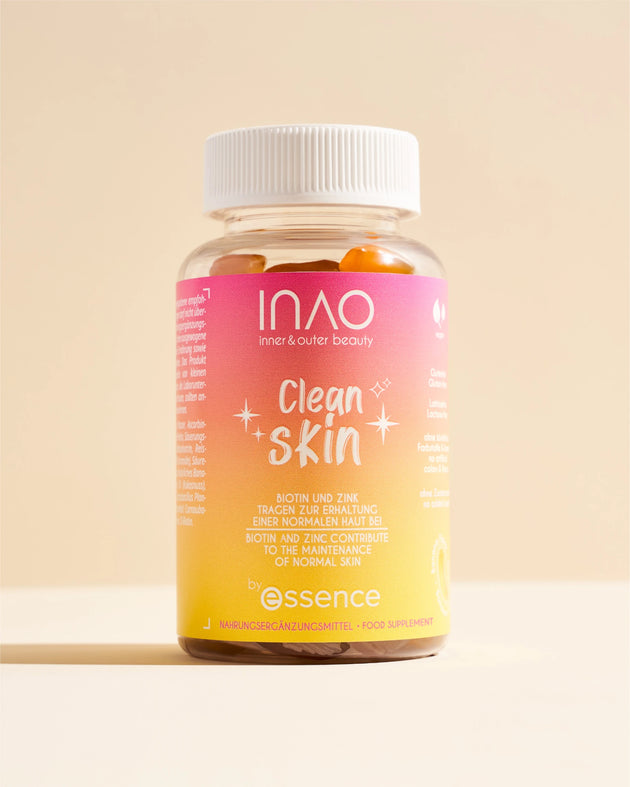 Eine Flasche INAO by essence Clean Skin Gummies mit weißem Verschluss enthält orangefarbene Gummibärchen. Das rosa-gelbe Etikett trägt weißen Text, und die Flasche steht auf einer beigen Fläche mit beigem Hintergrund.