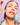 Eine Frau trägt INAO by essence Dewy Drop Deep Hydration Serum aus einer blauen Flasche mit einer Pipette auf ihre Wange auf, vor einem hellvioletten Hintergrund mit Kacheln. Sie trägt natürliches Make-up und ein leichtes Oberteil, das dessen feuchtigkeitsspendende Wirkung unterstreicht.