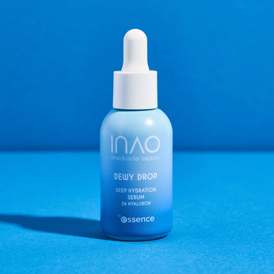 Eine blaue Flasche INAO by essence Dewy Drop Deep Hydration Serum mit weißem Tropfverschluss steht aufrecht auf blauem Untergrund. Das Etikett hebt 2 % Hyaluronsäure hervor, die Ihrer Haut einen tiefen Feuchtigkeitsschub verleiht.