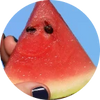 Eine Hand mit dunkel lackierten Nägeln hält ein Stück Wassermelone vor blauem Hintergrund. Zwei Wassermelonenkerne sind so angeordnet, dass sie wie Augen aussehen und der Frucht ein gesichtsähnliches Aussehen verleihen.