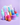 Die 24-Stunden-Hydro-Gel-Creme Hydra Soul 24h von INAO by essence in pastellfarbenen Tiegeln mit Regenbogenmotiven, angereichert mit Glycerin, steht auf einer violetten Fläche neben Sprays und Tropfern, im Hintergrund ist ein wiederverwendbarer Beutel mit Farbverlauf zu sehen.