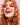 Eine lächelnde Person mit lockigem blondem Haar und leuchtend pinkem Lidschatten hält vor einem zartrosa Hintergrund ein rotes Rosenblatt zwischen den Zähnen – ein strahlender Look, der oft durch Hair YEAH! Gummies von INAO von essence unterstützt wird.