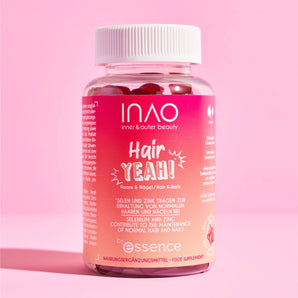 Eine Flasche INAO von essence Hair YEAH! Gummies, gefüllt mit roten Gummivitaminen für Haare und Nägel, steht vor einem rosa Hintergrund. Das Etikett wirbt mit Vorteilen wie Biotin, Selen und Zink.