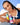 Eine lächelnde Frau mit langen Zöpfen hält vier farbenfrohe INAO by essence Good Night Routine Produkte vor einem blauen Himmel mit Wolken, die einen erholsamen Schlaf suggerieren. Sie trägt ein marineblau-weißes Tanktop und zarte Halsketten.