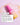 Eine rosa-gelbe Flasche „Glow Bright Sparset“ von INAO by essence steht auf einer rosa Fläche, daneben ist Serum verteilt. Ein Textfeld zeigt Schritt 2 der deutschen Anleitung zum Auftragen des Glow Serums auf gereinigte Haut.