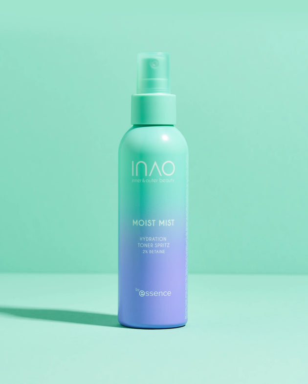 Ein blaugrüner und lavendelfarbener INAO von essence Moist Mist Hydration Toner Spritz steht aufrecht auf einer pastellfarbenen Oberfläche und hebt die feuchtigkeitsspendende Kraft von Betain vor einem minimalistischen Hintergrund in Blaugrün und Mintgrün hervor.