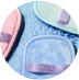 Drei pastellfarbene Badehandschuhe – Rosa, Lavendel und Hellgrün – jeweils mit einem farblich passenden Riemen, sind auf einer weichen, blau strukturierten Oberfläche angeordnet.