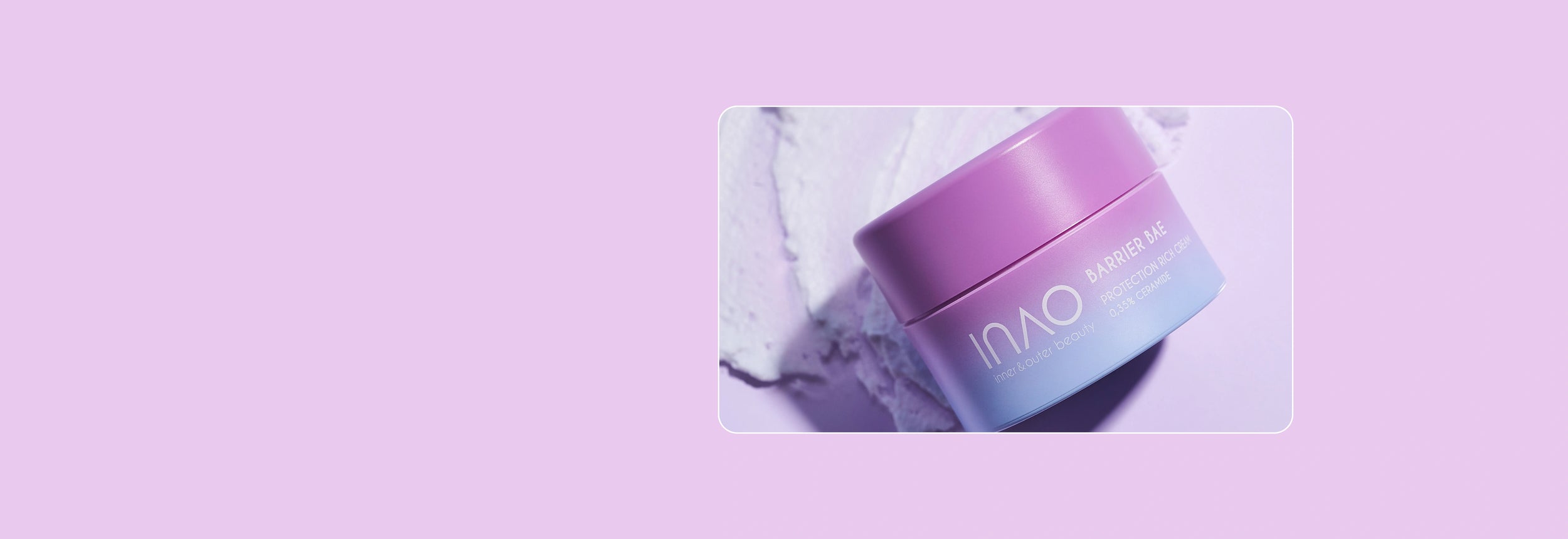 Eine lächelnde Frau mit Creme im Gesicht hält ein Glas Hautpflegeprodukt an ihre Wange und steht vor einer hellvioletten gefliesten Wand.