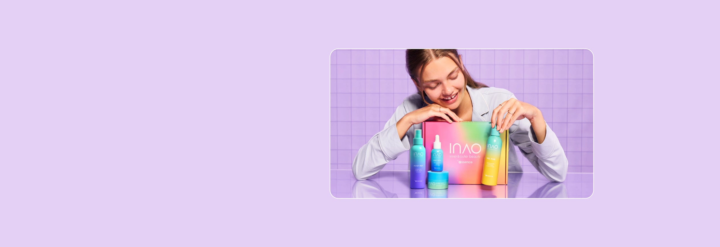 Eine lächelnde Frau mit glattem, dunklem Haar hält drei bunte Hautpflegeprodukte in ihren Händen und trägt einen flauschigen blau-violetten Pullover vor einem schlichten, hellen Hintergrund.