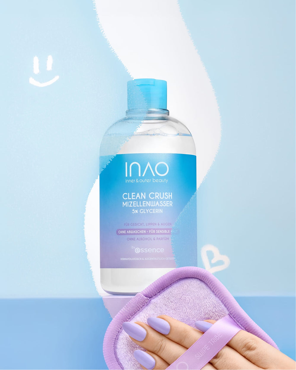 Eine Hand mit pastellvioletten Fingernägeln hält ein zartviolettes Tuch neben einer Flasche INAO Clean Crush Mizellenwasser in Reisegröße – ideal für empfindliche Haut – auf einer blauen Fläche, auf deren Hintergrund ein gekritzeltes Smiley-Gesicht und ein Herz zu sehen sind.
