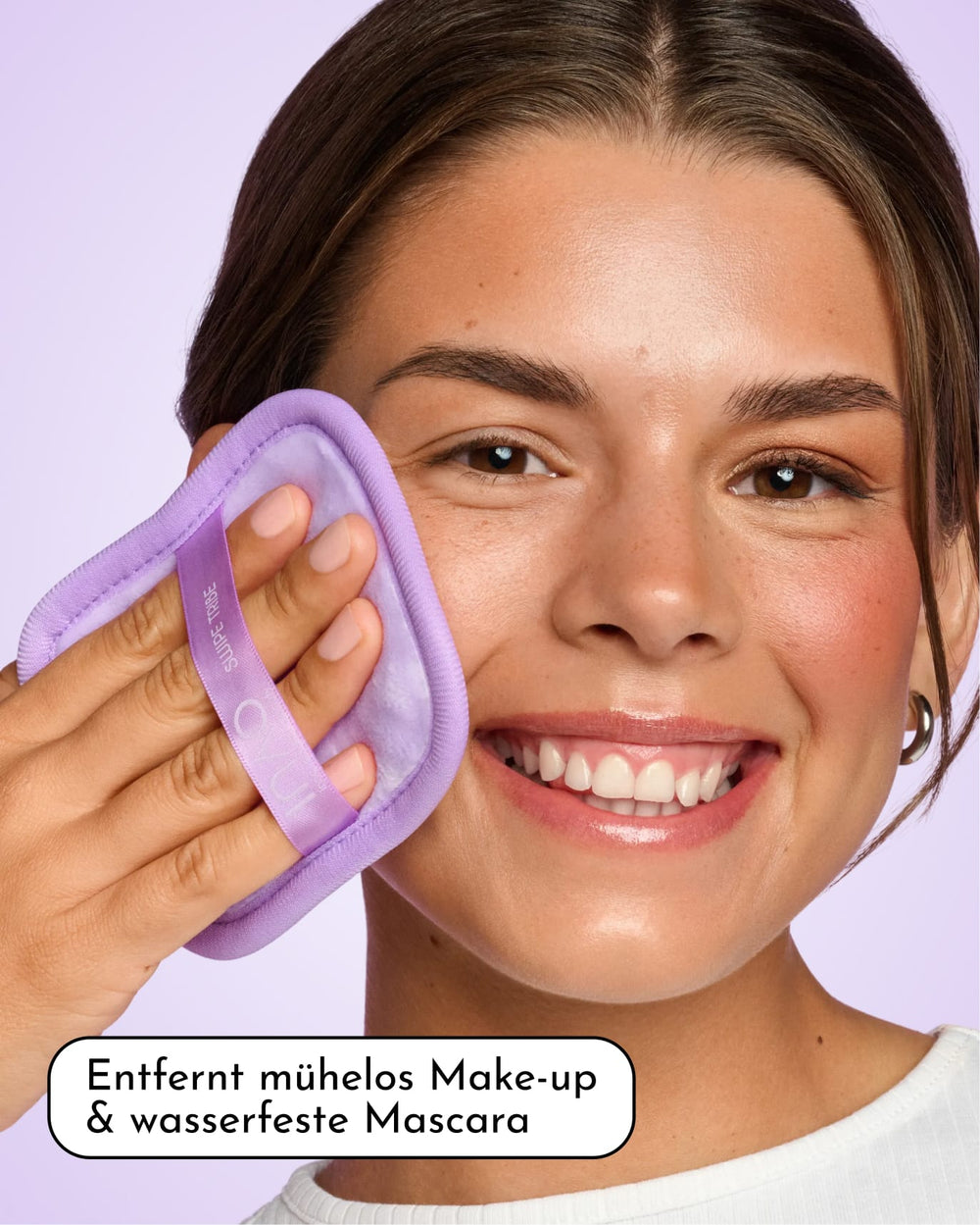 Eine lächelnde Person trägt das INAO Clean Crush Mizellenwasser in Reisegröße mit einem violetten Wattepad auf ihre Wange auf. Der deutsche Text lautet: „Entfernt mühelos Make-up & wasserfeste Mascara.“ Dieser sanfte Reiniger ist perfekt für empfindliche Haut geeignet und entfernt Make-up und wasserfeste Mascara, ohne die Haut zu reizen.