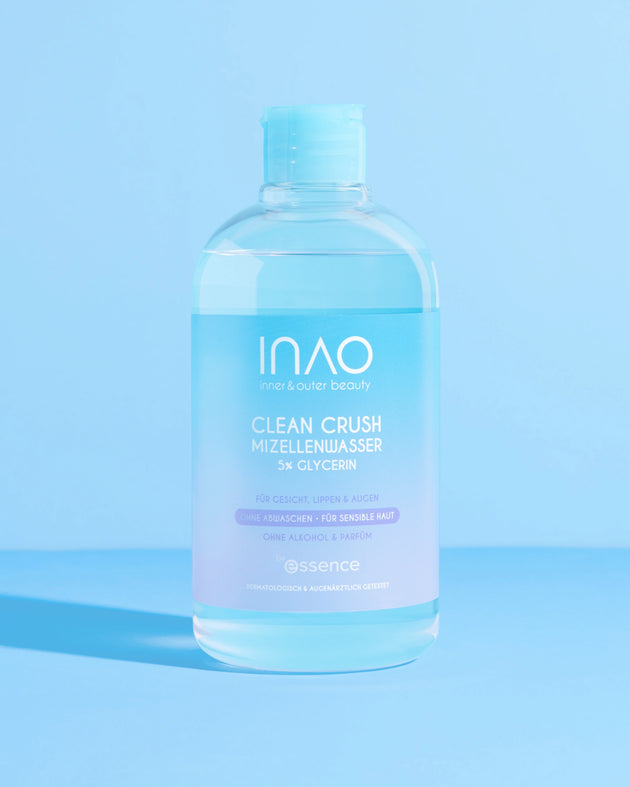 Eine klare Flasche INAO by essence Clean Crush Mizellenwasser mit 5% Glycerin steht auf einer hellblauen Fläche vor einem passenden blauen Hintergrund. Auf dem weiß-blauen Etikett steht lila Text, der die Vorteile des Produkts hervorhebt.
