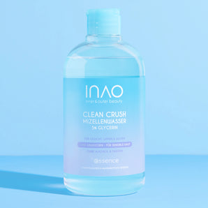 Eine klare Flasche INAO by essence Clean Crush Mizellenwasser mit 5% Glycerin steht auf einer hellblauen Fläche vor einem passenden blauen Hintergrund. Auf dem weiß-blauen Etikett steht lila Text, der die Vorteile des Produkts hervorhebt.