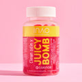 Eine durchsichtige Plastikflasche mit der Aufschrift „Clean Skin x JUICY BOMB Gummies“ von INAO by essence, gefüllt mit leuchtend rosa Gummibärchen, reich an Biotin und Zink, steht auf einer rosa Fläche mit rosa Hintergrund. Das Etikett hat gelb-rosa Text und einen weißen Verschluss.
