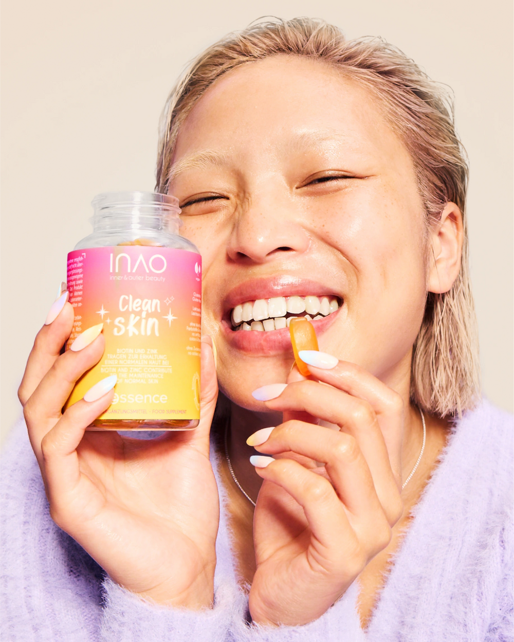 Eine lächelnde Person mit hellblondem Haar hält eine geöffnete Dose INAO by essence Clean Skin Gummies in der einen Hand und ein Gummibärchen in der anderen, bereit zum Verzehr. Sie trägt einen fliederfarbenen Pullover und hat manikürte Nägel.