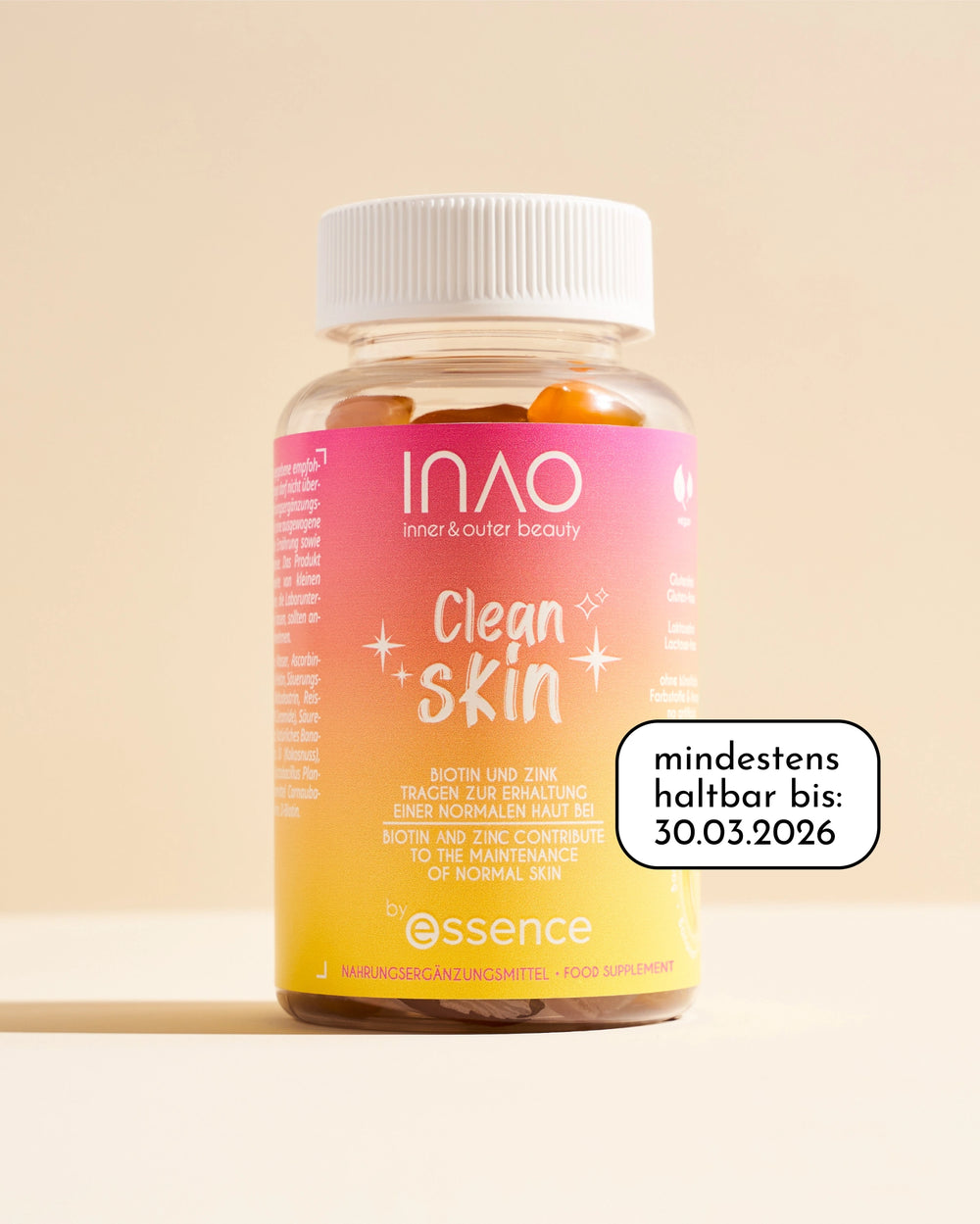 Eine Flasche INAO by essence Clean Skin Gummies, ein veganes Nahrungsergänzungsmittel mit Biotin und Zink, ist auf einem beigen Hintergrund abgebildet. Auf einem Aufkleber steht "mindestens haltbar bis: 30.03.2026.