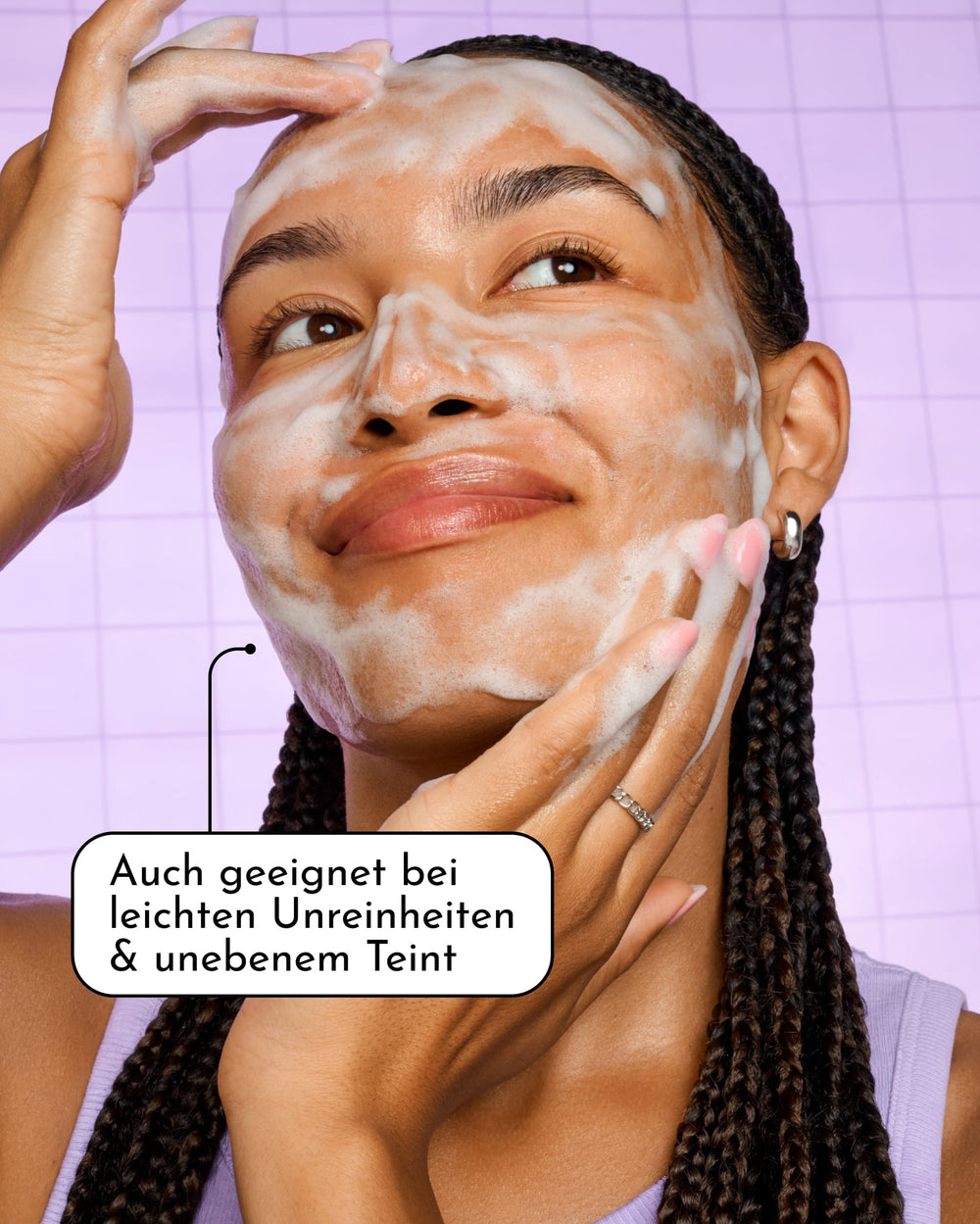 Eine Frau mit geflochtenen Haaren massiert sich sanft den schäumenden INAO Feel Pure Exfoliation Cleanser in Reisegröße ins Gesicht und lächelt dabei leicht. Der deutsche Text lautet: „Auch geeignet bei leichten Unreinheiten & unebenem Teint.“ Die Szene hebt die milde tägliche Reinigung vor einem hellvioletten Fliesenhintergrund hervor.
