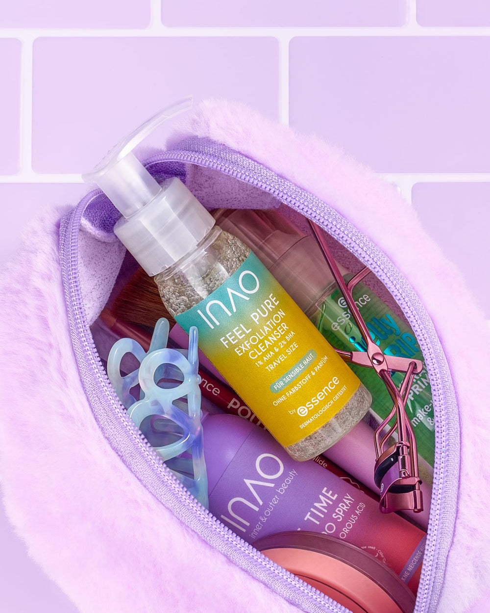 Eine flauschige, lavendelfarbene Kosmetiktasche, gefüllt mit Hautpflege- und Schönheitsprodukten, darunter eine Pumpflasche in Reisegröße mit dem „INAO Feel Pure Exfoliation Cleanser“ für die sanfte tägliche Reinigung, Essenzprodukte, eine Schere, eine Wimpernzange und weitere Kosmetikartikel, vor einem Hintergrund aus violetten Fliesen.