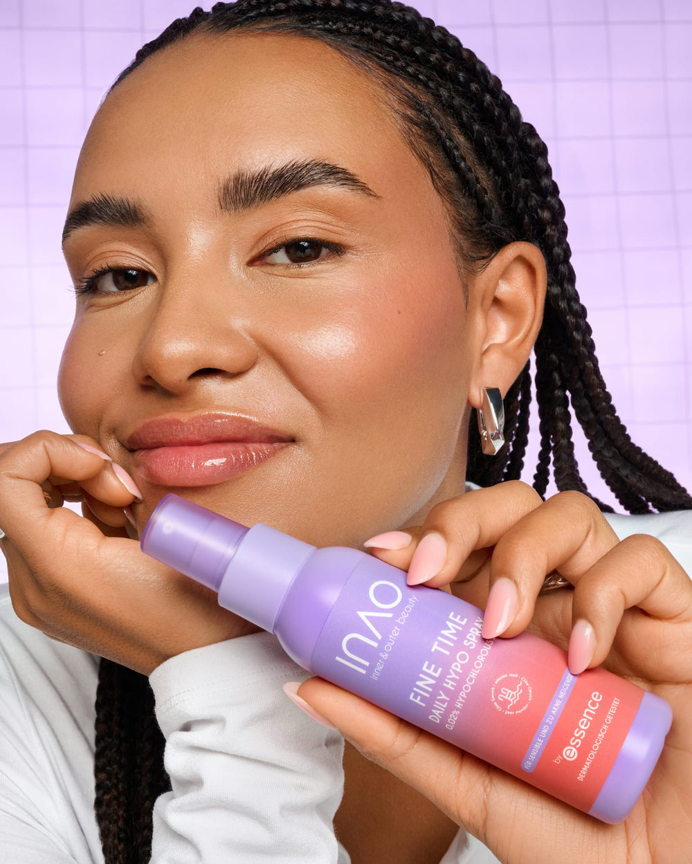 Eine Frau mit geflochtenen Haaren lächelt sanft, während sie vor einem violetten Rasterhintergrund eine violette Flasche mit der Aufschrift „Fine Time Daily Hypo Spray“ von INAO by essence in der Hand hält und so dieses Gesichtsspray hervorhebt, das die Haut beruhigt.