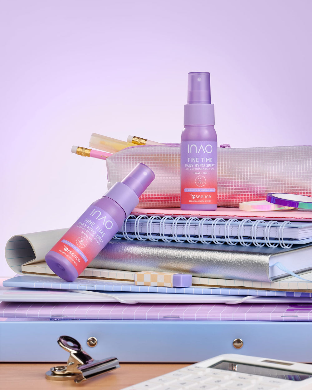 Zwei violette Flaschen des „Essence Fine Time“-Serums stehen auf einem Stapel aus Notizbüchern, Ordnern und Federmäppchen, umgeben von Bleistiften, Textmarkern und pastellfarbenem Schreibzeug. Ein Gesichtsspray wie das „Fine Time Daily Hypo Spray“ von INAO by essence würde diese beruhigende Kulisse perfekt ergänzen, da es die Haut vor dem sanften Farbverlauf von Hellviolett bis Rosa beruhigt.