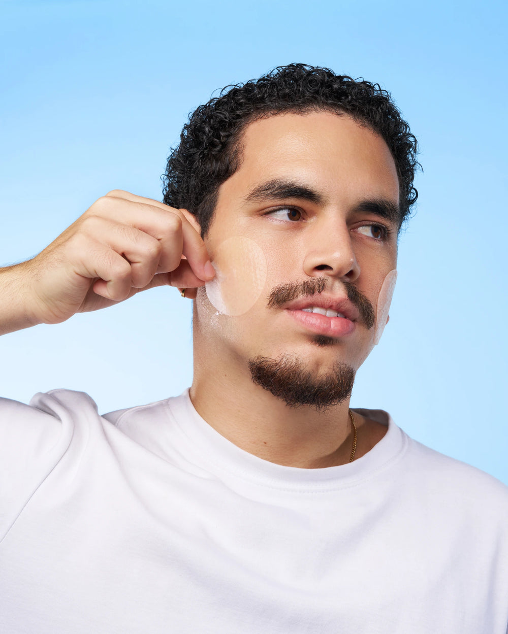 Ein Mann mit kurzen lockigen Haaren und einem Ziegenbart, der ein weißes Hemd trägt, schält vor einem hellblauen Hintergrund ein INAO by essence Hydra Soul 24h Hydra Jelly Toner Pad von seiner Wange.