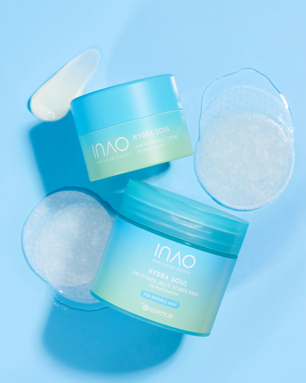 Zwei blau-grüne Tiegel der INAO by essence "Hydra Soul 24h Hydra Jelly Toner Pads" stehen auf einem hellblauen Hintergrund, der mit einer Gel-Creme und einem Abstrich in der Nähe gezeigt wird und die intensive Feuchtigkeitspflege hervorhebt.