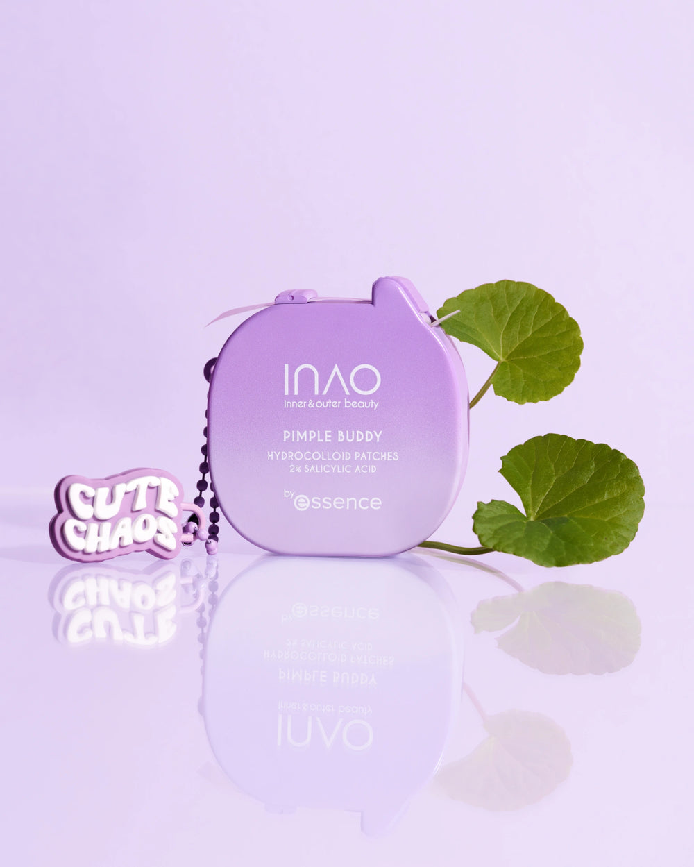Ein lavendelfarbenes Döschen mit der Aufschrift "Pimple Buddy Hydrocolloid Patches" von INAO by essence liegt auf einer zartvioletten Fläche mit zwei grünen Blättern und einem "CUTE CHAOS" Schlüsselanhänger. Der Hintergrund ist ebenfalls in einem hellen Lila-Ton gehalten.