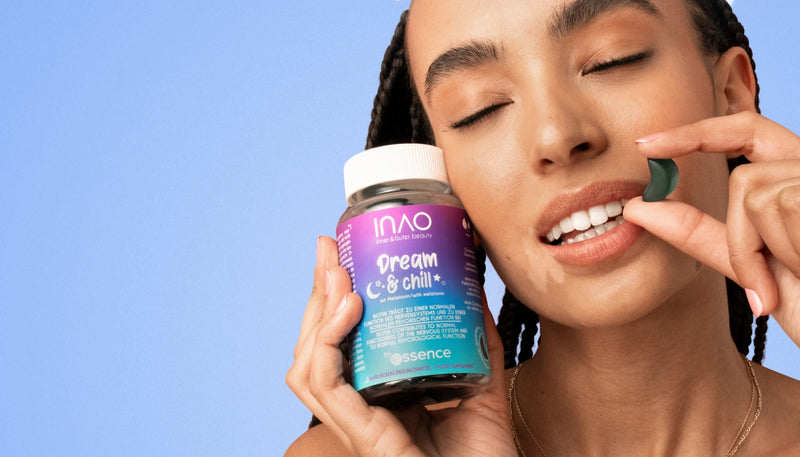 Eine lächelnde Frau mit geflochtenem Haar hält in einer Hand ein Glas mit der Aufschrift „Dream & Chill“-Gummibärchen und dicht an ihrem Mund ein grünes Gummibärchen mit hellblauem Hintergrund und dem Logo „by essence“ in der Ecke.