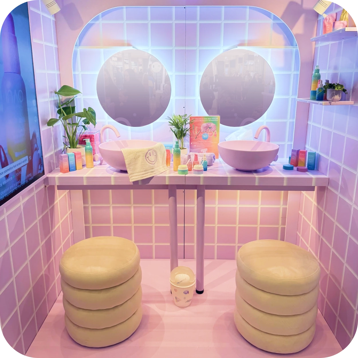 Pastellfarbener Schminktisch mit zwei runden Spiegeln, Waschbecken, Pflanzen, Hautpflegeprodukten und zwei gepolsterten Hockern. Wände und Boden sind in zartem Lavendel gefliest und schaffen so eine helle, verspielte Atmosphäre.