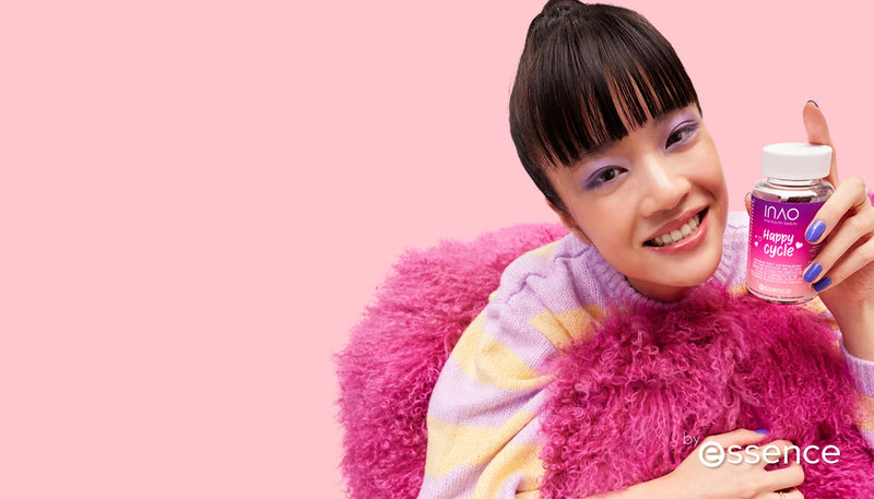 Eine lächelnde Person mit Pony und lila Augen-Make-up hält eine Flasche mit der Aufschrift „Happy Cycle“. Sie trägt einen pastellfarbenen Pullover und einen rosa flauschigen Schal. Der Hintergrund ist hellrosa, mit dem „Essence“-Logo in der Ecke.