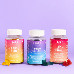 Drei bunte Flaschen aus dem INAO by essence Supplement Lover Sparset - "Clean Skin Gummies", "Dream & Chill" und "Hair YEAH! Gummies" - stehen nebeneinander auf einem lilafarbenen Hintergrund, vor dem jeweils passende Gummibärchen verstreut sind.